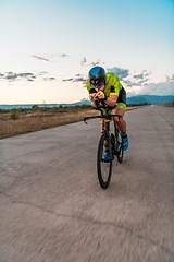 Image showing Triathlete riding his bicycle during sunset, preparing for a marathon. The warm colors of the sky provide a beautiful backdrop for his determined and focused effort.