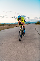 Image showing Triathlete riding his bicycle during sunset, preparing for a marathon. The warm colors of the sky provide a beautiful backdrop for his determined and focused effort.