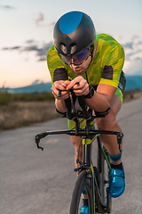 Image showing Triathlete riding his bicycle during sunset, preparing for a marathon. The warm colors of the sky provide a beautiful backdrop for his determined and focused effort.