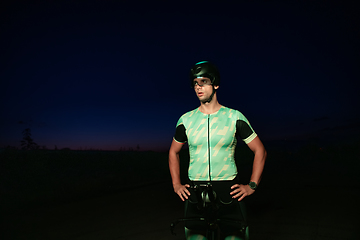 Image showing A triathlete resting on the road after a tough bike ride in the dark night, leaning on his bike in complete exhaustion