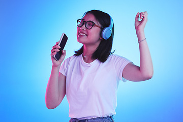 Image showing Singing, music and a woman with a phone on a blue background for a podcast, audio or fun. Happy, freedom and an Asian girl with headphones and a mobile app for radio, karaoke or sound on a backdrop