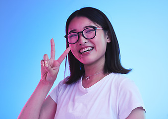 Image showing Peace sign, smile and show with portrait of woman in studio for emoji, like and social media. Happy, support and motivation with face of Asian person on blue background for v symbol, gen z and icon