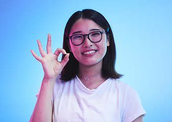 Image showing Woman, okay hands or sign for education success, certified and excellence in tshirt and a blue, studio background. Happy young, asian person in glasses, portrait and yes or support emoji for learning