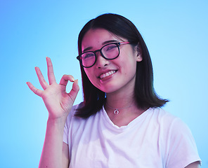 Image showing Woman, okay hands or sign for learning success, certified and excellence in tshirt and a blue, studio background. Happy young, asian person in glasses, portrait and yes or support emoji for education