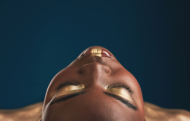 Image showing Beauty, gold makeup and art on face of black woman with creative gold aesthetics isolated in a studio blue background. Artistic, cosmetic and young person eyes closed for fantasy with color or paint