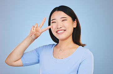 Image showing Peace, sign and portrait of woman with hand for emoji in studio blue background with gen z style, fashion or happiness. Face, smile and Asian model with gesture, expression