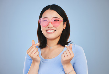 Image showing Happy asian woman, portrait and sunglasses with love hands, sign or gesture against a blue studio background. Female person smile in happiness with loving emoji, icon or symbol for valentines day