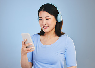 Image showing Woman, phone and headphones for listening, audio streaming and social media on a blue background. Young asian person with music subscription, electronics and mobile chat or web networking in studio