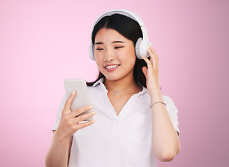 Image showing Headphones, smile and woman with a phone in a studio listening to music, playlist or album for entertainment. Happy, calm and Asian female model streaming a song or radio isolated by pink background.