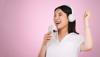 Image showing Headphones, singing and woman with a phone in a studio listening to music, playlist or radio. Happy, smile and young Asian female model doing karaoke with a cellphone mic to a song by pink background