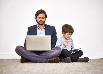 Image showing Boys love their digital toys. A father and son sitting on the floor against a wall with a laptop and digital tablet.