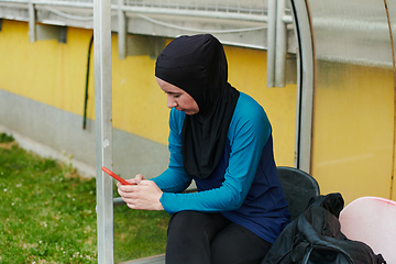 Image showing Muslim sports fitness woman dressed in hijab and dark clothes outdoors in green nature park using mobile phone chatting