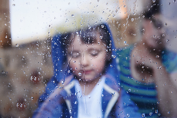 Image showing Rainy days with nothing to do.... A little boy looking downwards behind a rain-streaked window while his mother sits in the background.