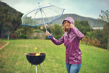 Image showing Only the brave barbeque in the rain. Shot of a woman holiding an umbrella while trying to barbeque in the rain.