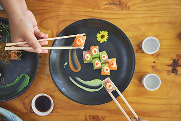 Image showing Chopsticks, hand and eating sushi and food at a restaurant for nutrition at table. Above hungry people with wooden sticks and soy sauce for dining, Japanese culture and cuisine with art on a plate