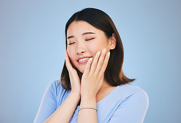 Image showing Face, beauty and skincare with an asian woman on a blue background for natural treatment or cosmetics. Smile, makeup and freedom with a young model touching her skin in satisfaction in a studio