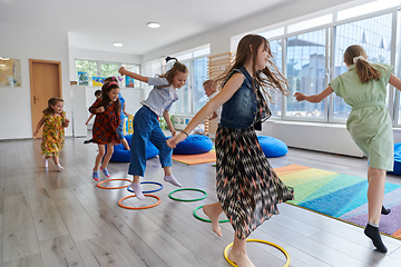 Image showing Small nursery school children with female teacher on floor indoors in classroom, doing exercise. Jumping over hula hoop circles track on the floor.