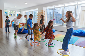 Image showing Small nursery school children with female teacher on floor indoors in classroom, doing exercise. Jumping over hula hoop circles track on the floor.