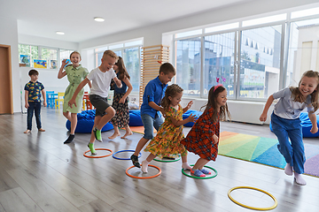 Image showing Small nursery school children with female teacher on floor indoors in classroom, doing exercise. Jumping over hula hoop circles track on the floor.