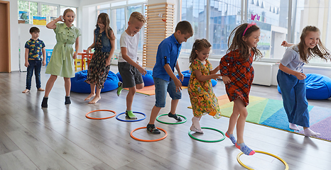 Image showing Small nursery school children with female teacher on floor indoors in classroom, doing exercise. Jumping over hula hoop circles track on the floor.