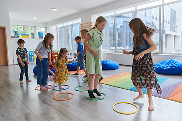 Image showing Small nursery school children with female teacher on floor indoors in classroom, doing exercise. Jumping over hula hoop circles track on the floor.