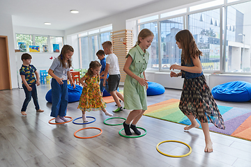 Image showing Small nursery school children with female teacher on floor indoors in classroom, doing exercise. Jumping over hula hoop circles track on the floor.