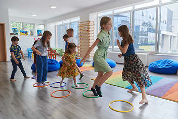 Image showing Small nursery school children with female teacher on floor indoors in classroom, doing exercise. Jumping over hula hoop circles track on the floor.