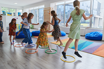 Image showing Small nursery school children with female teacher on floor indoors in classroom, doing exercise. Jumping over hula hoop circles track on the floor.