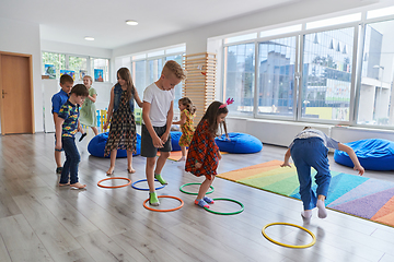 Image showing Small nursery school children with female teacher on floor indoors in classroom, doing exercise. Jumping over hula hoop circles track on the floor.