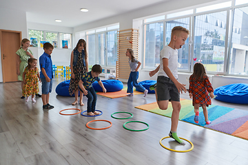 Image showing Small nursery school children with female teacher on floor indoors in classroom, doing exercise. Jumping over hula hoop circles track on the floor.