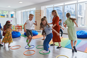 Image showing Small nursery school children with female teacher on floor indoors in classroom, doing exercise. Jumping over hula hoop circles track on the floor.