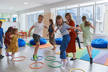 Image showing Small nursery school children with female teacher on floor indoors in classroom, doing exercise. Jumping over hula hoop circles track on the floor.