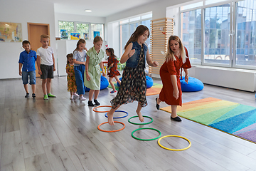 Image showing Small nursery school children with female teacher on floor indoors in classroom, doing exercise. Jumping over hula hoop circles track on the floor.