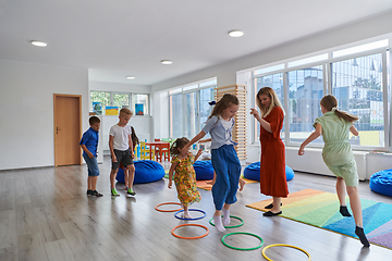 Image showing Small nursery school children with female teacher on floor indoors in classroom, doing exercise. Jumping over hula hoop circles track on the floor.