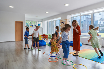 Image showing Small nursery school children with female teacher on floor indoors in classroom, doing exercise. Jumping over hula hoop circles track on the floor.