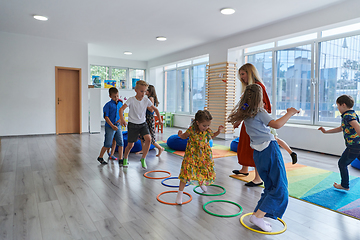 Image showing Small nursery school children with female teacher on floor indoors in classroom, doing exercise. Jumping over hula hoop circles track on the floor.