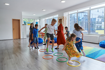 Image showing Small nursery school children with female teacher on floor indoors in classroom, doing exercise. Jumping over hula hoop circles track on the floor.