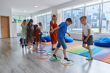 Image showing Small nursery school children with female teacher on floor indoors in classroom, doing exercise. Jumping over hula hoop circles track on the floor.