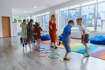 Image showing Small nursery school children with female teacher on floor indoors in classroom, doing exercise. Jumping over hula hoop circles track on the floor.