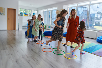 Image showing Small nursery school children with female teacher on floor indoors in classroom, doing exercise. Jumping over hula hoop circles track on the floor.