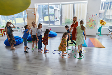 Image showing Small nursery school children with female teacher on floor indoors in classroom, doing exercise. Jumping over hula hoop circles track on the floor.