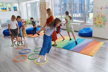 Image showing Small nursery school children with female teacher on floor indoors in classroom, doing exercise. Jumping over hula hoop circles track on the floor.