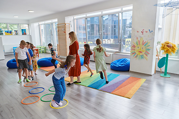 Image showing Small nursery school children with female teacher on floor indoors in classroom, doing exercise. Jumping over hula hoop circles track on the floor.