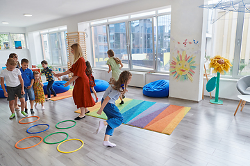 Image showing Small nursery school children with female teacher on floor indoors in classroom, doing exercise. Jumping over hula hoop circles track on the floor.