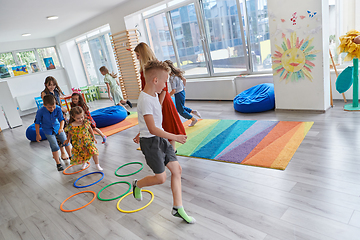 Image showing Small nursery school children with female teacher on floor indoors in classroom, doing exercise. Jumping over hula hoop circles track on the floor.