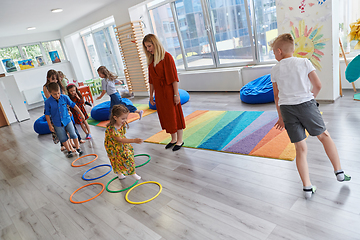 Image showing Small nursery school children with female teacher on floor indoors in classroom, doing exercise. Jumping over hula hoop circles track on the floor.