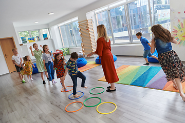 Image showing Small nursery school children with female teacher on floor indoors in classroom, doing exercise. Jumping over hula hoop circles track on the floor.