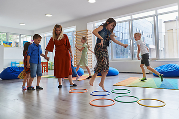 Image showing Small nursery school children with female teacher on floor indoors in classroom, doing exercise. Jumping over hula hoop circles track on the floor.