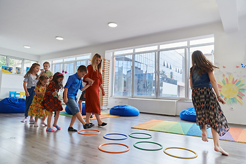 Image showing Small nursery school children with female teacher on floor indoors in classroom, doing exercise. Jumping over hula hoop circles track on the floor.