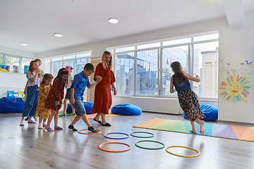 Image showing Small nursery school children with female teacher on floor indoors in classroom, doing exercise. Jumping over hula hoop circles track on the floor.